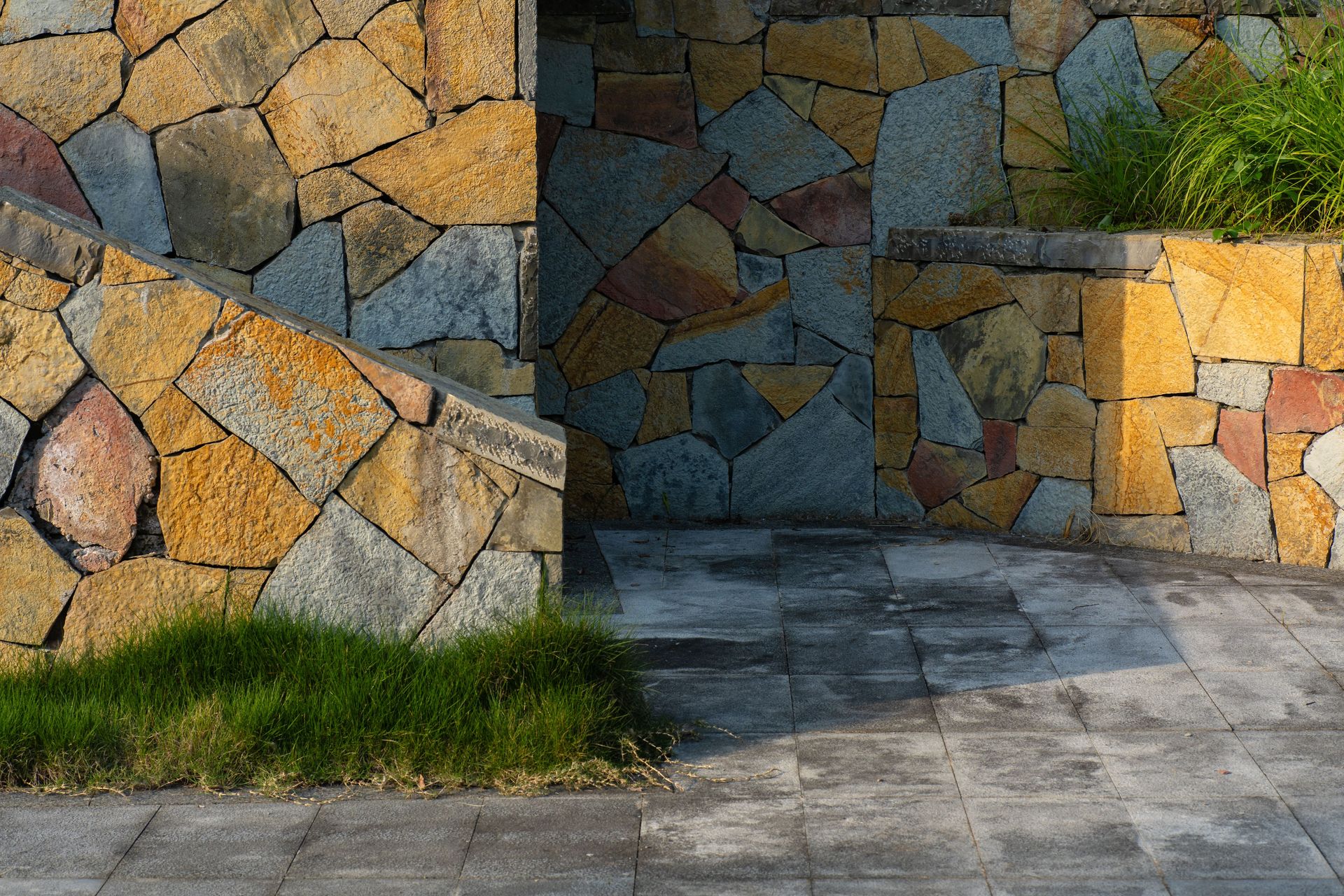 Close-up of a colorful stone wall with paved ground and lush grass in sunlight.